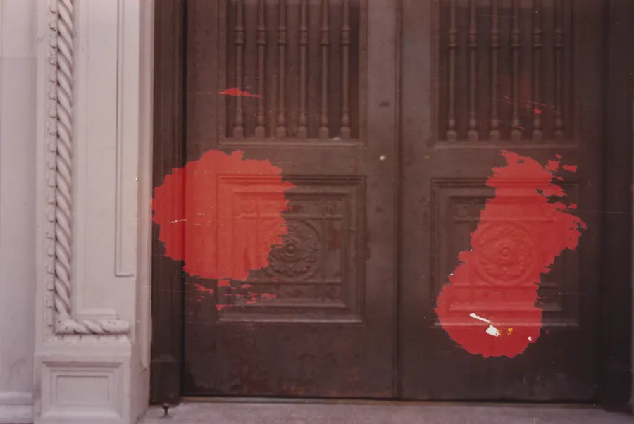 Photograph of dark carved wooden double doors beside a white door frame, with two patches of red paint dabbed over the panels