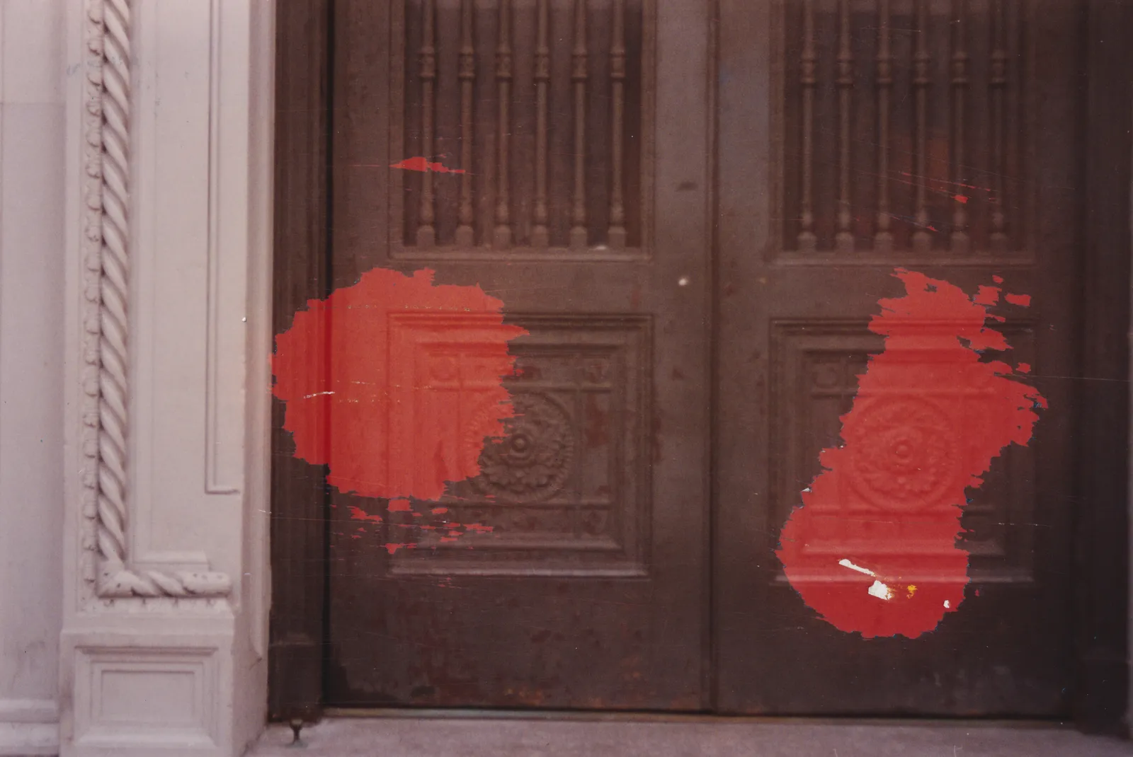 Photograph of dark carved wooden double doors beside a white door frame, with two patches of red paint dabbed over the panels