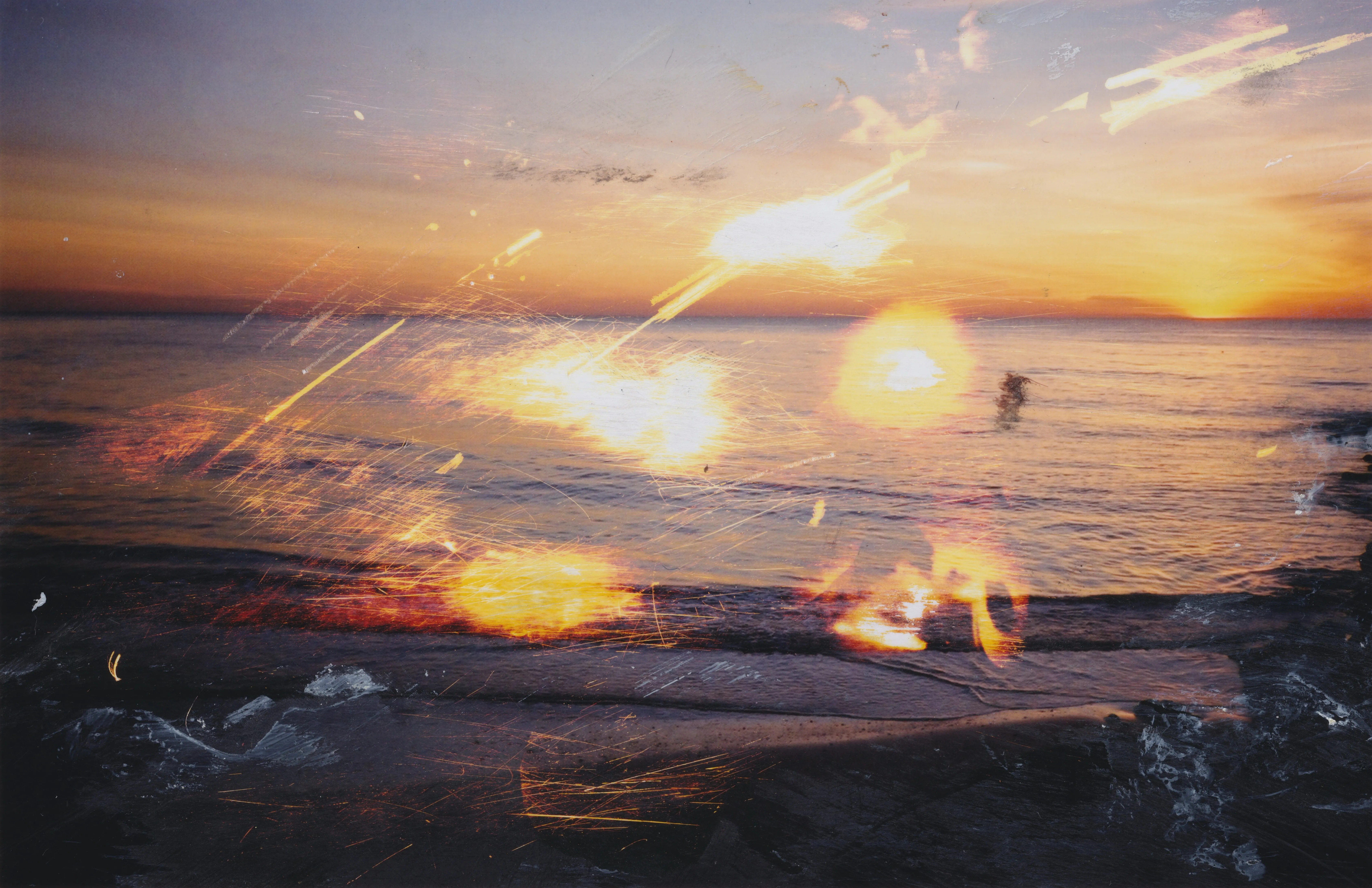 Photograph of a sunset over the sea from a dark shore, with scratched streaks of light, glowing orange spots and dabs of white paint