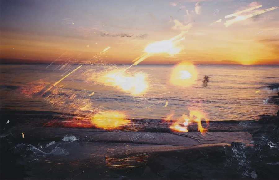 Photograph of a sunset over the sea from a dark shore, with scratched streaks of light, glowing orange spots and dabs of white paint
