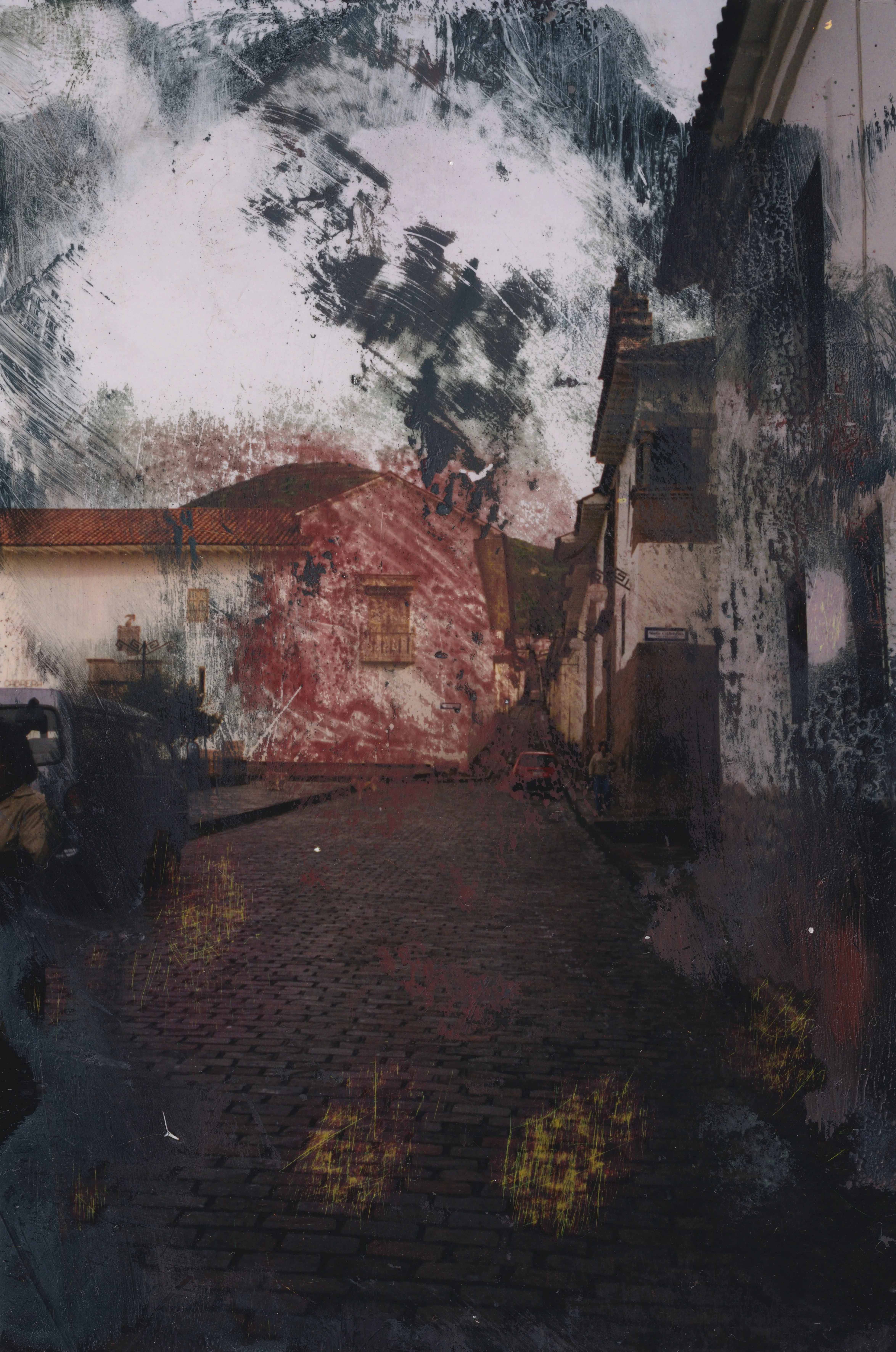 Photograph of a cobbled village street between white houses, the sky smeared with dark paint, a red wash on a far wall and faint yellow rings on the cobbles