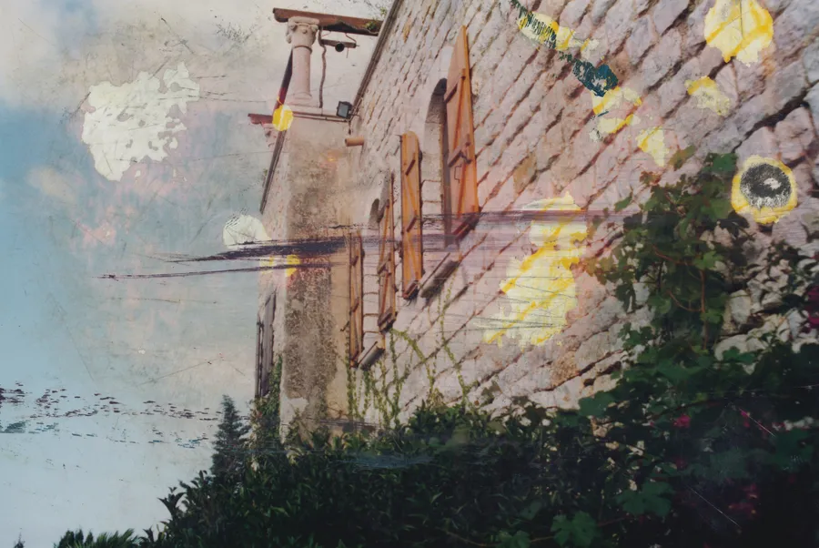 Photograph looking up at a stone house with wooden shutters against a pale sky, dark trees below, with white and yellow paint marks and thin dark streaks